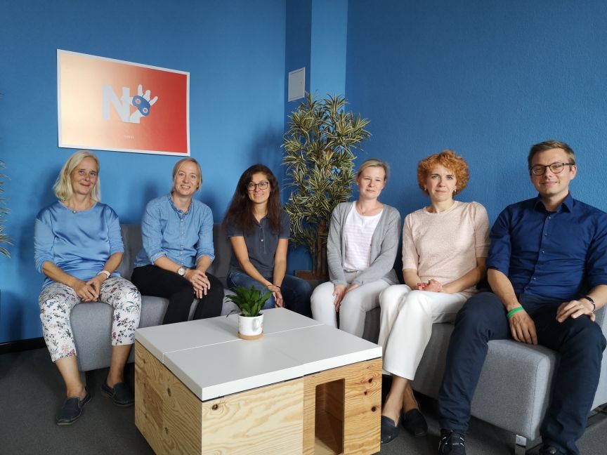 Sechs Personen sitzen auf grauen Sofas in einem blauen Büroraum vor einem Logo-Schild an der Wand.