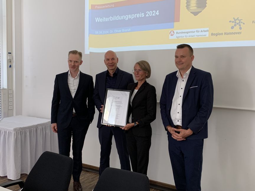 Vier Personen stehen vor einer Präsentation „Weiterbildungspreis 2024". Zwei halten gemeinsam eine gerahmte Urkunde.
