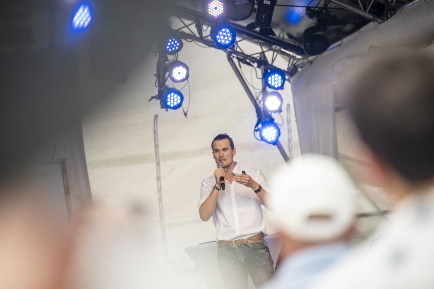 Mann in weißem Hemd spricht mit Mikrofon auf einer Bühne mit blauen LED-Scheinwerfern vor Publikum.