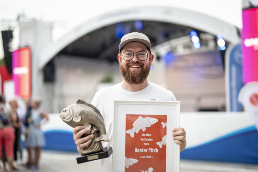 Mann mit Brille und Bart hält Fisch-Trophäe und Urkunde „Bester Pitch 500 Euro" vor einer Bühne.