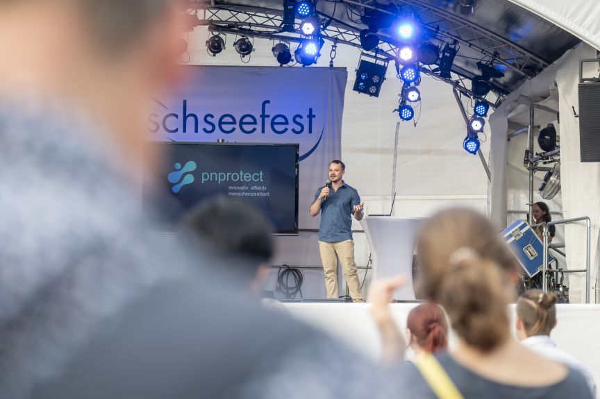Mann spricht mit Mikrofon auf Bühne beim Schseefest, hinter ihm ein Banner mit "pnprotect"-Logo, Publikum im Vordergrund.