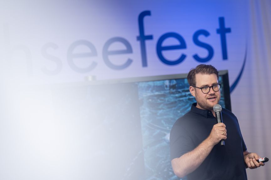 Mann mit Brille hält Mikrofon und spricht vor einem Banner mit der Aufschrift „seefest" auf einer Bühne.