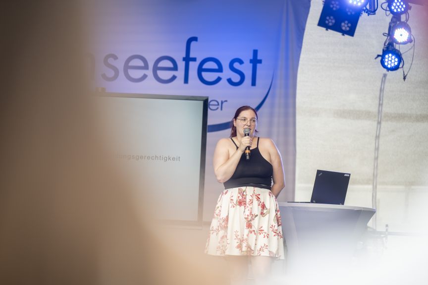 Frau spricht mit Mikrofon auf einer Bühne vor einem Banner mit der Aufschrift „seefest". Im Hintergrund Beleuchtung und Laptop.