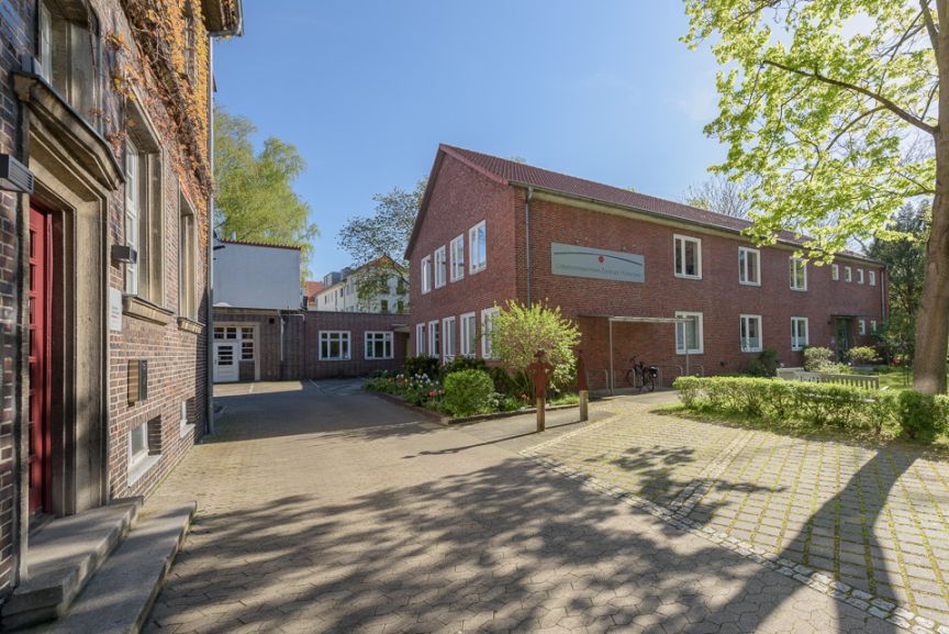 Gepflasterter Innenhof mit roten Backsteingebäuden, Grünflächen und Bäumen bei sonnigem Wetter.