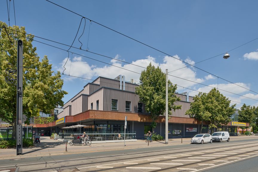 Moderne Gebäude mit Holzfassade an einer Stadtstraße mit Straßenbahngleisen, Bäumen und parkenden Autos.