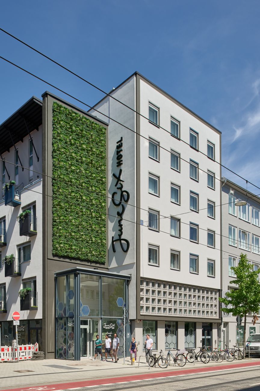 Modernes Stadtgebäude mit begrünter Fassadenwand, Glaseingang, Fahrrädern und Fußgängern auf dem Gehweg.