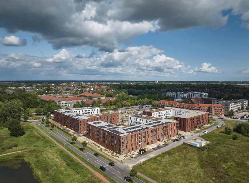 Luftaufnahme eines Stadtviertels mit roten Backsteingebäuden, Solaranlagen auf Dächern, Straßen und grüner Umgebung.