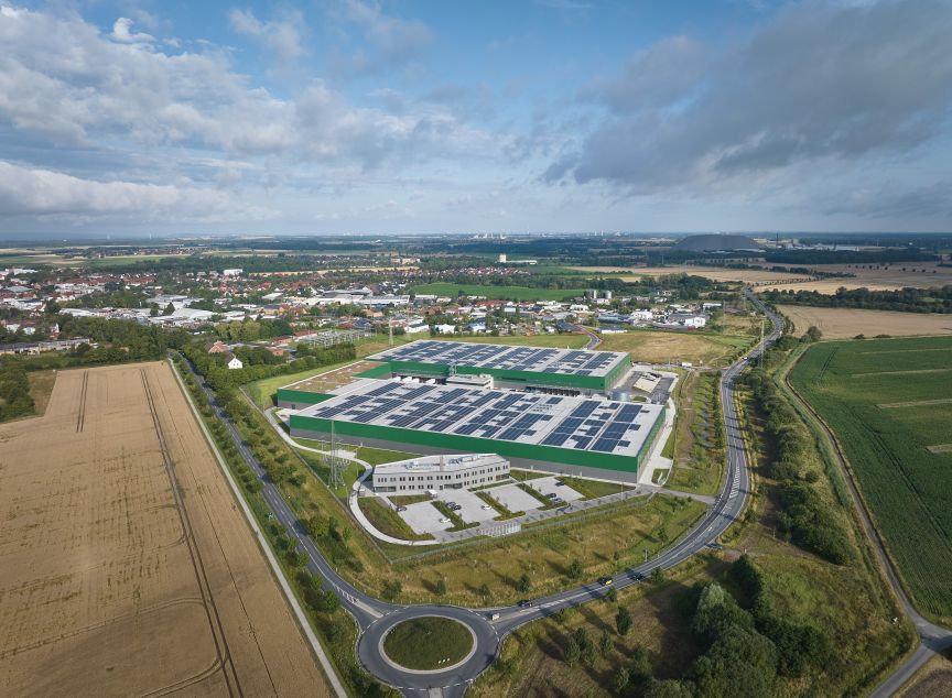 Luftaufnahme eines großen Industriegebäudes mit Solarpanelen auf dem Dach, umgeben von Feldern und einer Kleinstadt.
