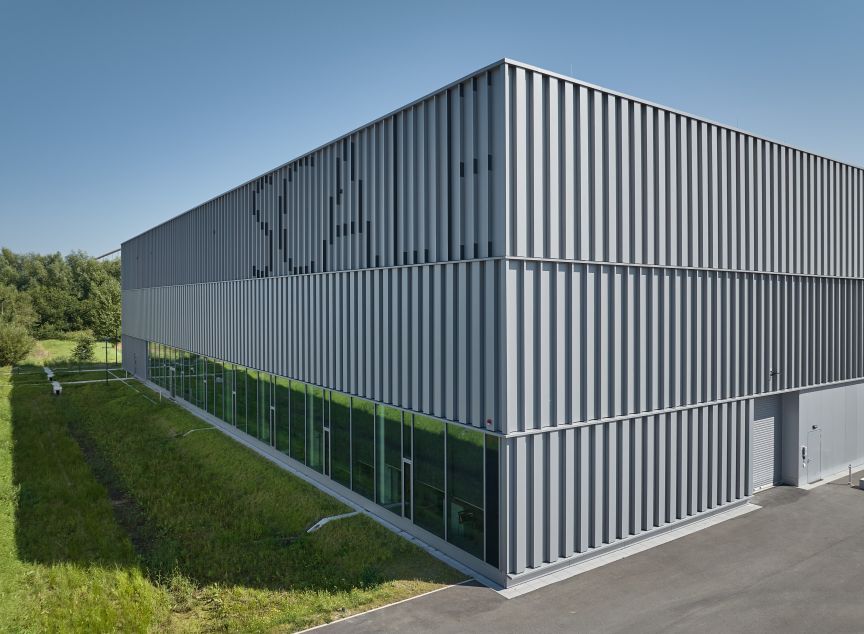 Modernes Industriegebäude mit grauer Metallfassade, vertikalen Lamellen und Glasfront im Erdgeschoss, umgeben von Grünfläche.