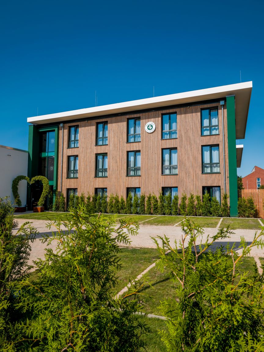 Modernes dreigeschossiges Gebäude mit Holzfassade und grünen Akzenten, umgeben von Büschen und Bäumen bei blauem Himmel.