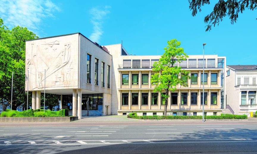 Modernes Verwaltungsgebäude mit Reliefschmuck, mehrstöckiger Fassade, Bäumen und breiter Straße bei blauem Himmel.