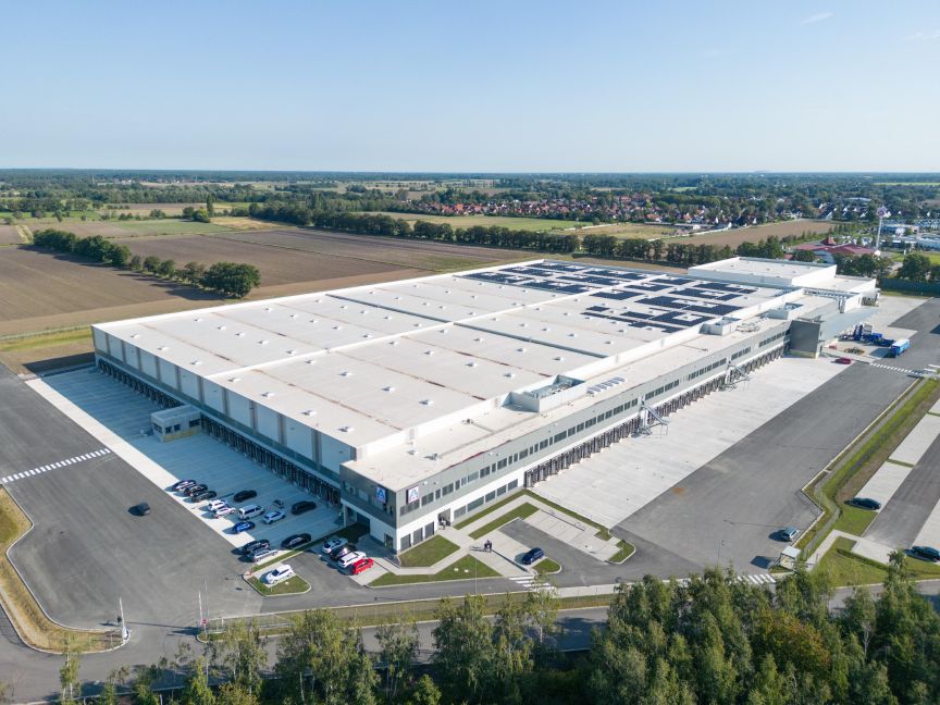 Luftaufnahme eines großen weißen Industriegebäudes mit Solarpanelen auf dem Dach, Parkplatz und umliegenden Feldern.