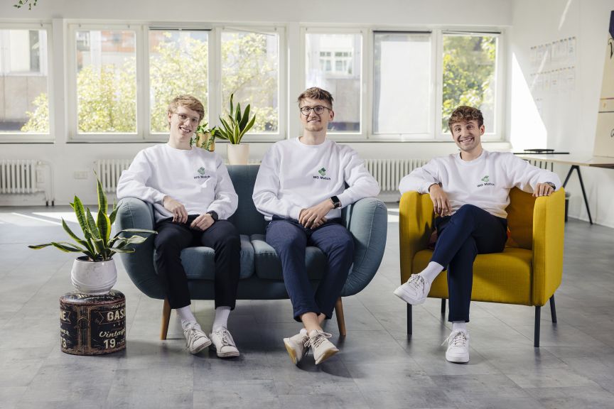 Drei junge Männer in weißen Sweatshirts sitzen lächelnd auf Sesseln in einem hellen Büroraum.