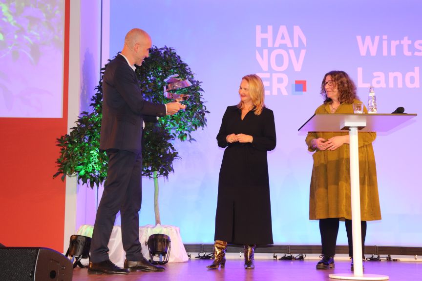 Drei Personen auf einer Bühne vor einem "Hannover"-Banner, eine Person hält eine Auszeichnung.