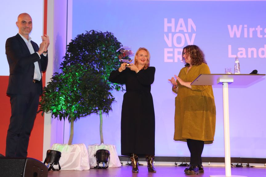 Drei Personen auf einer Bühne, eine hält eine Glasauszeichnung hoch. Im Hintergrund ein "Hannover"-Banner.