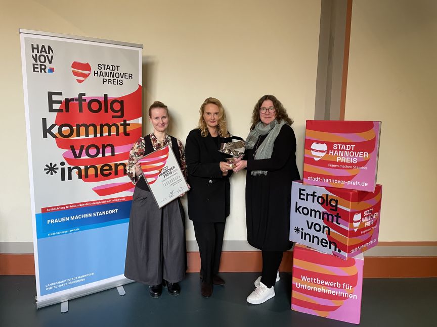 Drei Frauen stehen vor einem Roll-up-Banner des Stadt Hannover Preises. Eine hält ein Zertifikat, eine andere eine Trophäe.