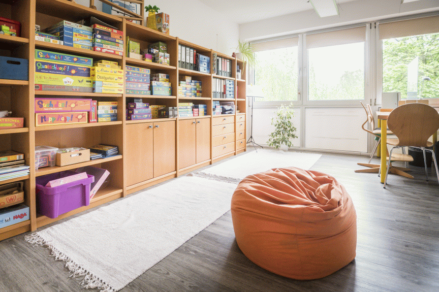 Heller Raum mit großem Bücherregal voller Spiele, orangefarbenem Sitzsack auf Holzboden, weißem Teppich und Fensterfront.