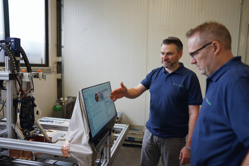 Zwei Männer in dunkelblauen Poloshirts betrachten einen Computermonitor neben einer Industriemaschine.