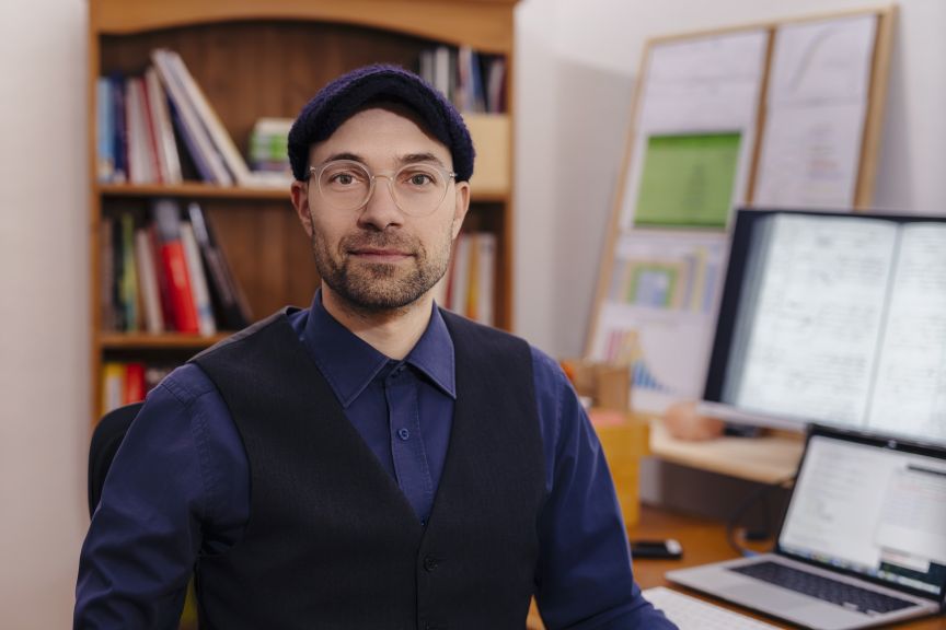 Mann mit Brille und blauer Mütze in schwarzer Weste sitzt vor Bücherregal und Laptop im Büro.