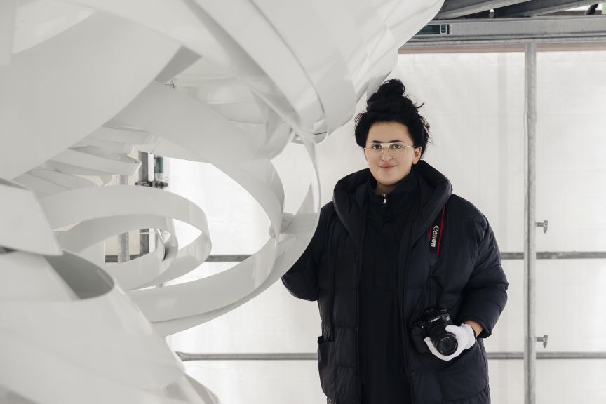 Frau mit schwarzer Daunenjacke und Canon-Kamera steht vor weißer spiralförmiger Skulptur in hellem Raum.