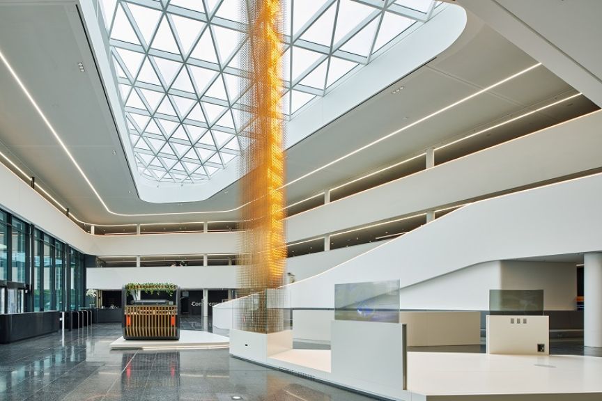 Modernes Atrium mit Glasdach, goldenem Hängeobjekt, weißen Treppen und Empfangsbereich auf poliertem Steinboden.