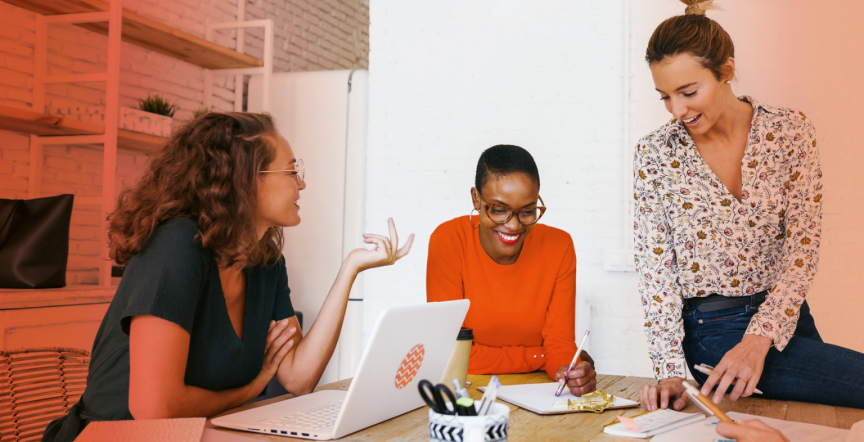 Drei Frauen arbeiten gemeinsam an einem Tisch mit Laptop und Notizen in einem Büro.