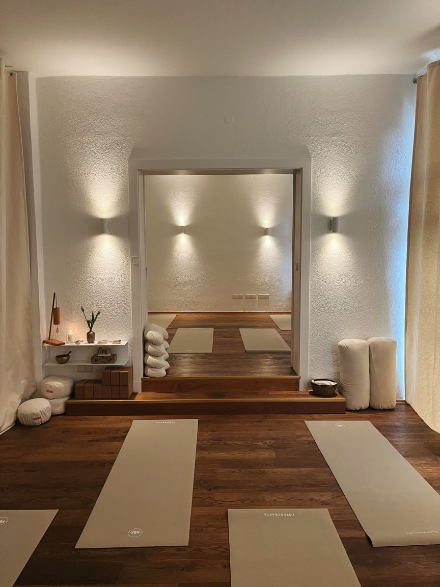 Heller Yogaraum mit vier beigen Matten auf Holzboden, Wandleuchten, Meditationskissen und einem Altar mit Kerzen.