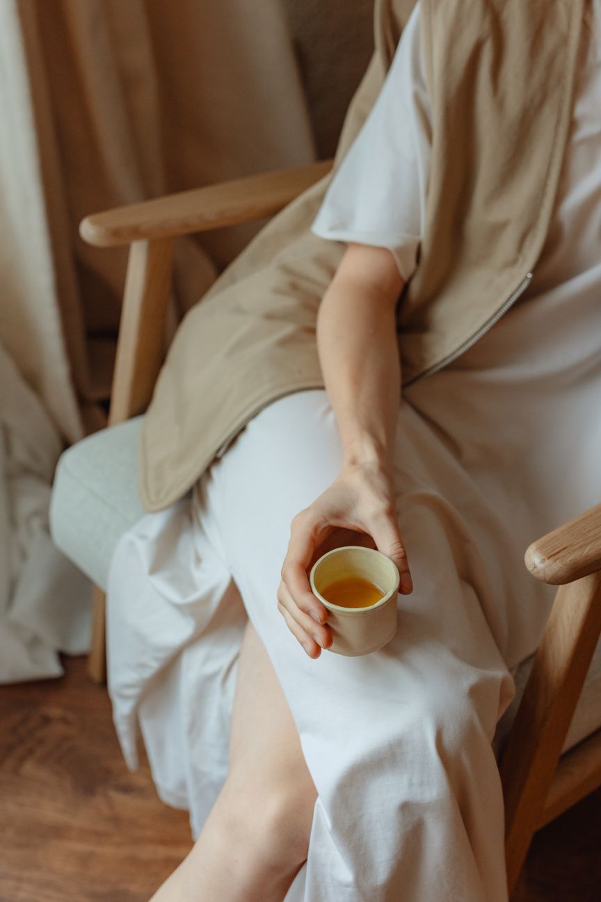 Person in weißem Kleid sitzt auf Holzstuhl und hält eine kleine Tasse mit gelbem Getränk in der Hand.
