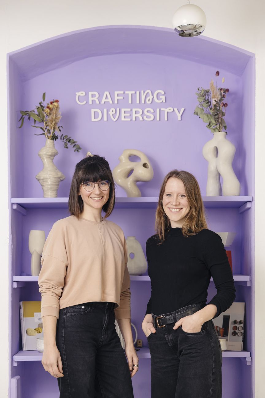 Zwei Frauen stehen vor einem lila Regal mit der Aufschrift „Crafting Diversity" und dekorierten Vasen.