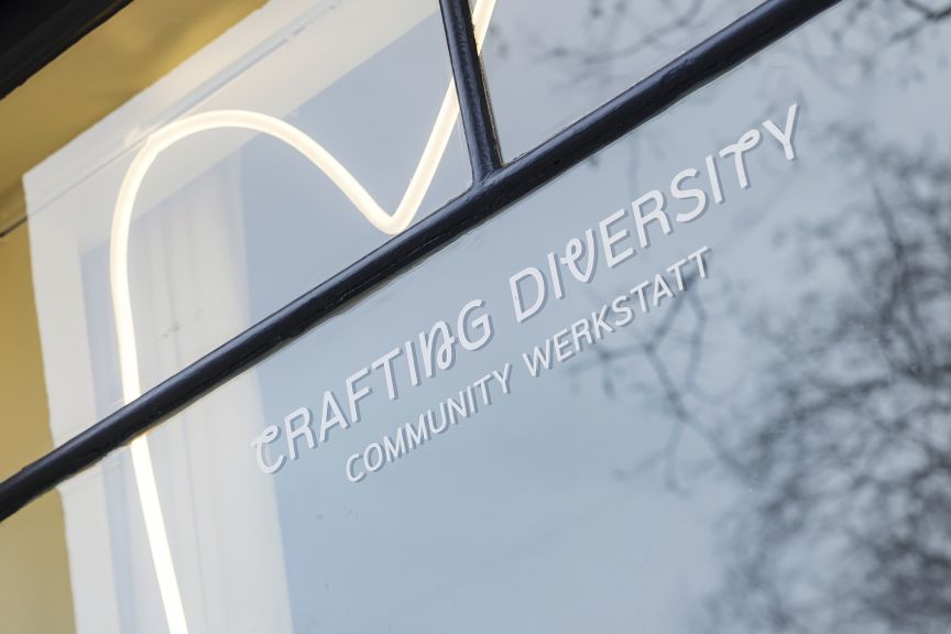 Glasscheibe mit weißer Aufschrift „Crafting Diversity – Community Werkstatt" und einem Neonlicht-Logo dahinter.