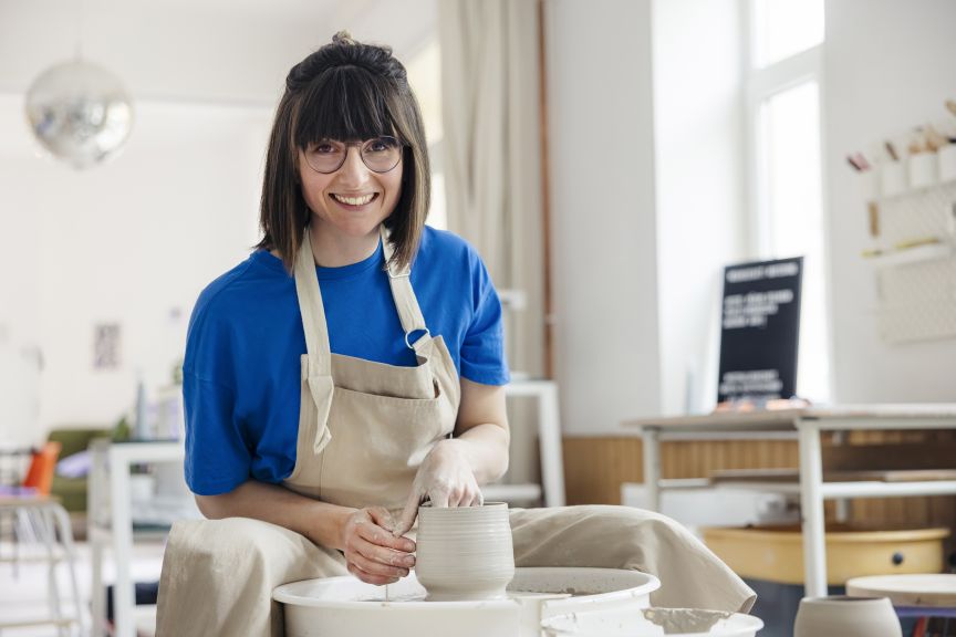 Frau mit Brille und Schürze formt Ton auf einer Töpferscheibe in einem hellen Atelier.