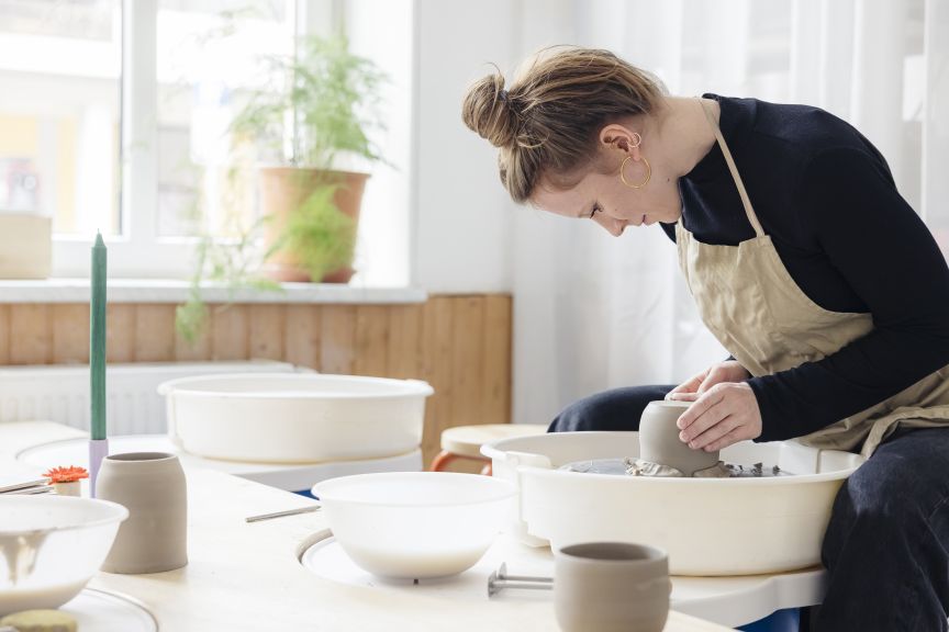 Frau in Schürze arbeitet konzentriert an einer Töpferscheibe in einem hellen Atelier mit fertigen Keramikstücken.