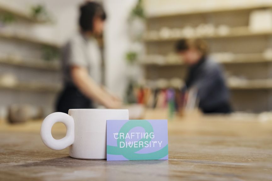 Visitenkarte „Crafting Diversity" lehnt an weißer Tasse auf Holztisch, im Hintergrund zwei Personen in einem Töpferstudio.