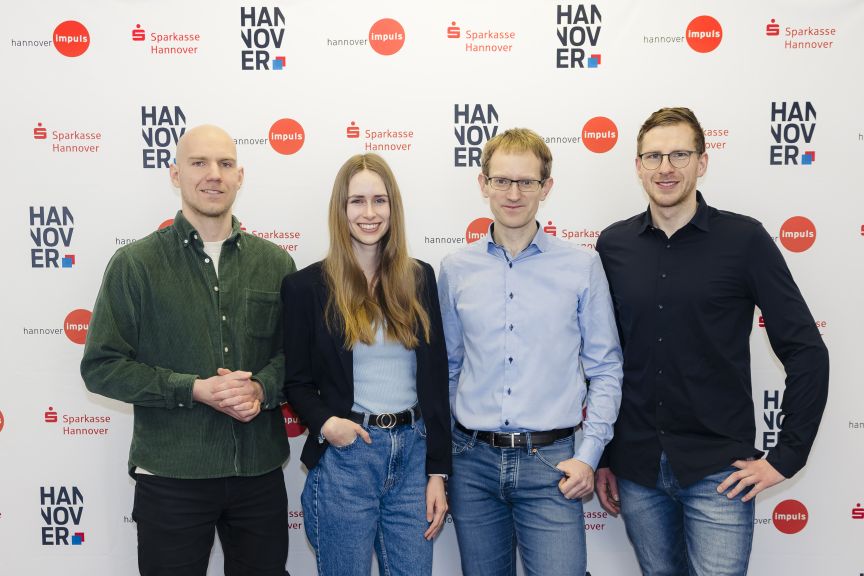 Vier Personen stehen lächelnd vor einer Sponsorenwand mit Logos von Sparkasse Hannover und Hannover Impuls.