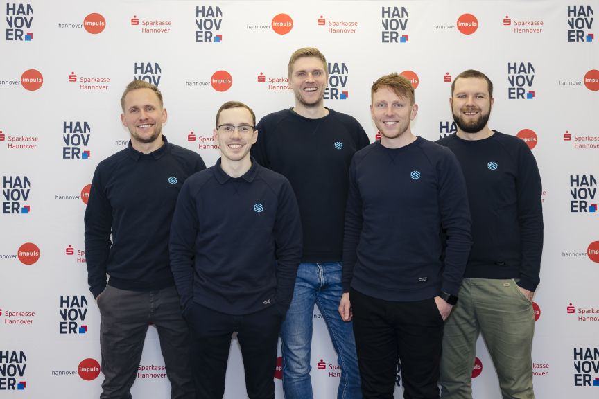 Fünf Männer in dunklen Sweatshirts posieren lächelnd vor einer Hannover-Impuls- und Sparkasse-Hannover-Sponsorenwand.