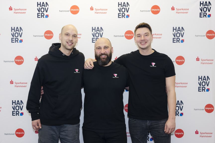 Drei Männer in schwarzen Shirts posieren lächelnd vor einem Hannover-Sparkasse-Werbebanner.