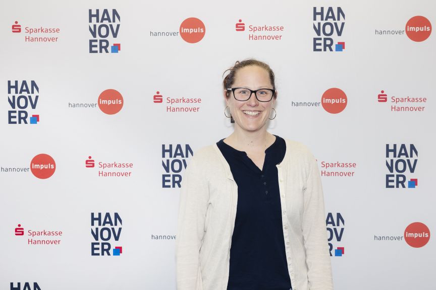 Frau mit Brille und hellen Haaren steht vor einer Sponsorenwand mit Logos von Sparkasse Hannover und hannover impuls.