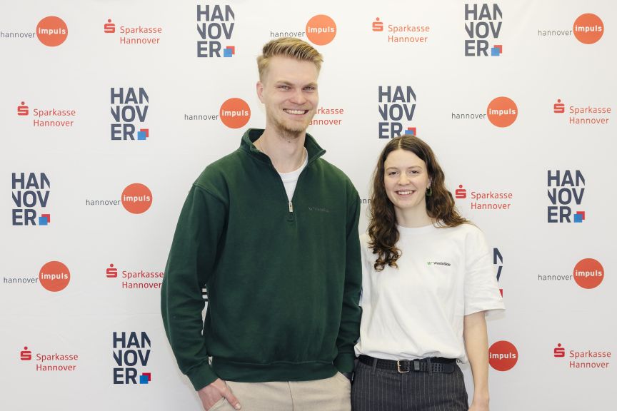 Zwei junge Personen stehen lächelnd vor einer Sponsorenwand mit Logos von Sparkasse Hannover und Hannover Impuls.