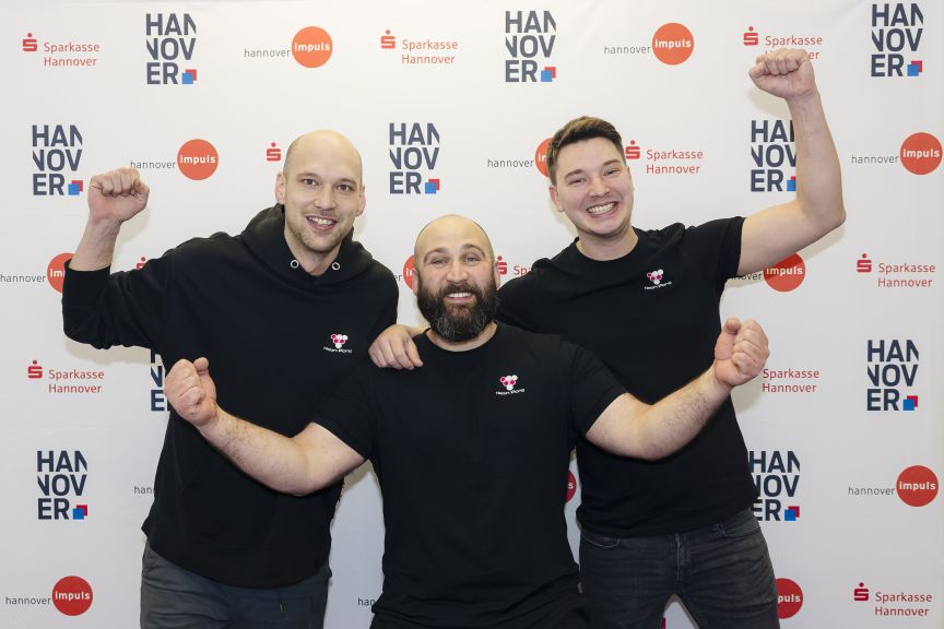 Drei Männer in schwarzen Hoodies posieren jubelnd vor einer Sparkasse Hannover Pressewall.