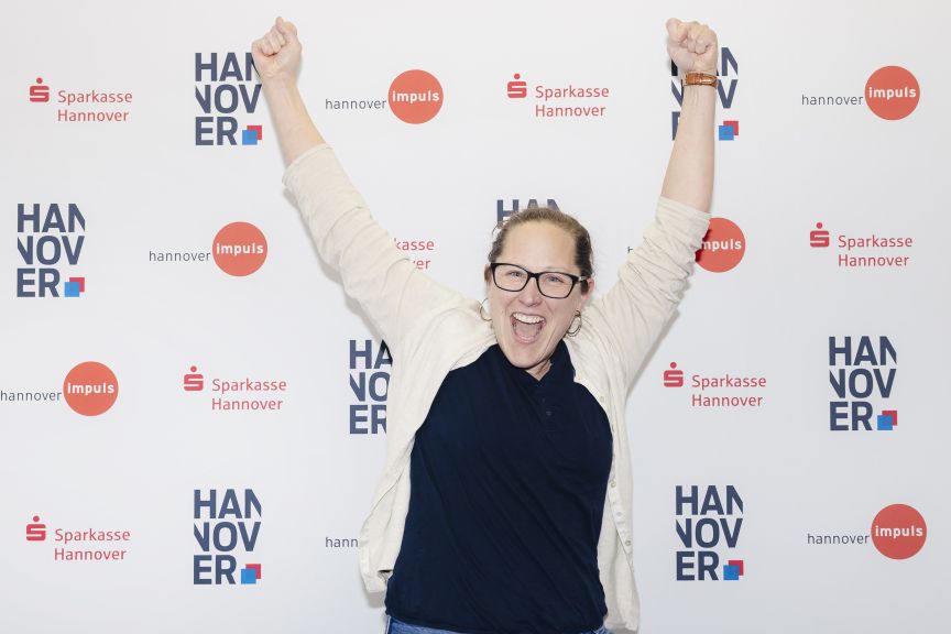 Frau mit erhobenen Fäusten und offenem Mund vor einer Sponsorenwand mit „Sparkasse Hannover" und „hannover impuls"-Logos.