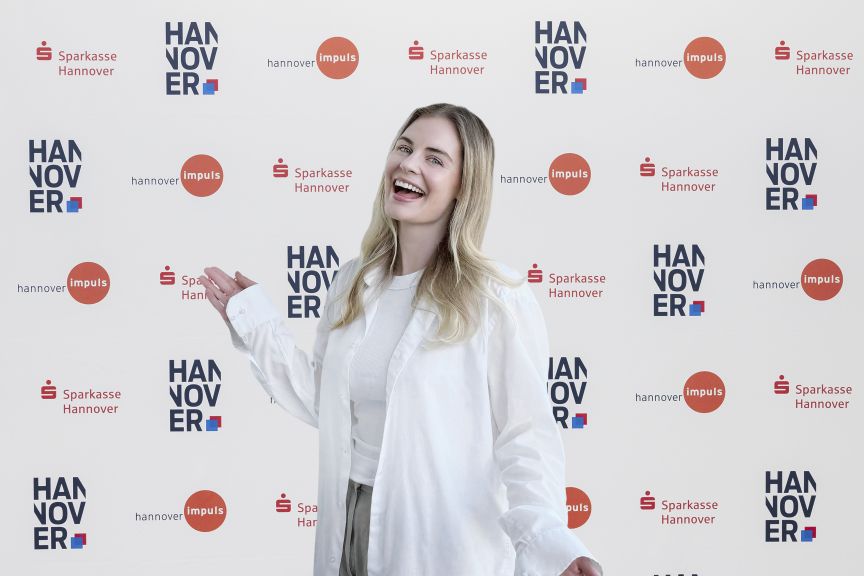 Lachende blonde Frau in weißem Hemd vor einer Sponsorenwand mit Logos von Sparkasse Hannover und Hannover Impuls.