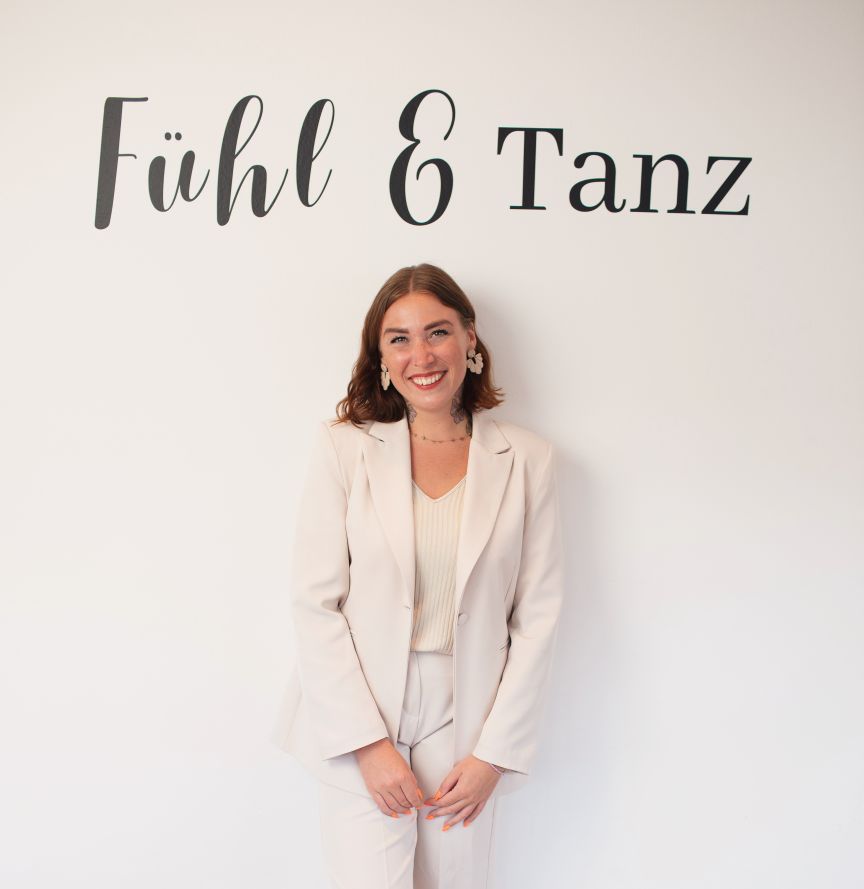 Junge Frau in beigem Hosenanzug lächelt vor weißer Wand mit der Aufschrift „Fühl & Tanz".