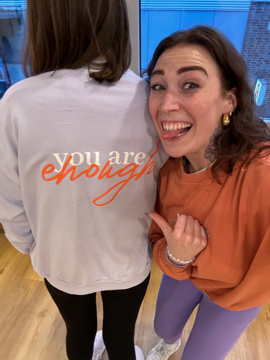 Zwei Frauen; eine trägt ein weißes Sweatshirt mit Aufschrift „you are enough", die andere lächelt und zeigt darauf.