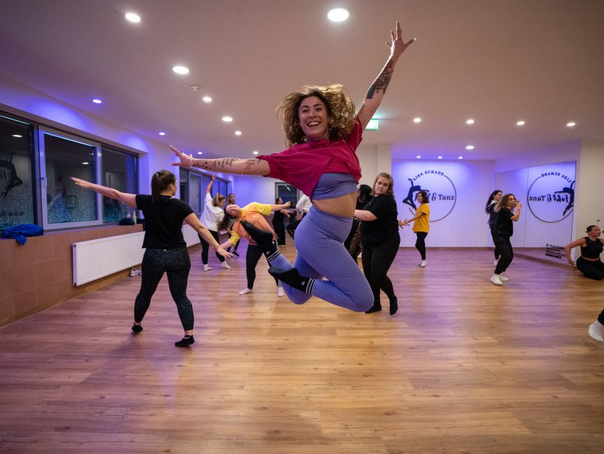 Eine Frau springt lächelnd in einem Tanzstudio in die Luft, im Hintergrund tanzen weitere Personen.