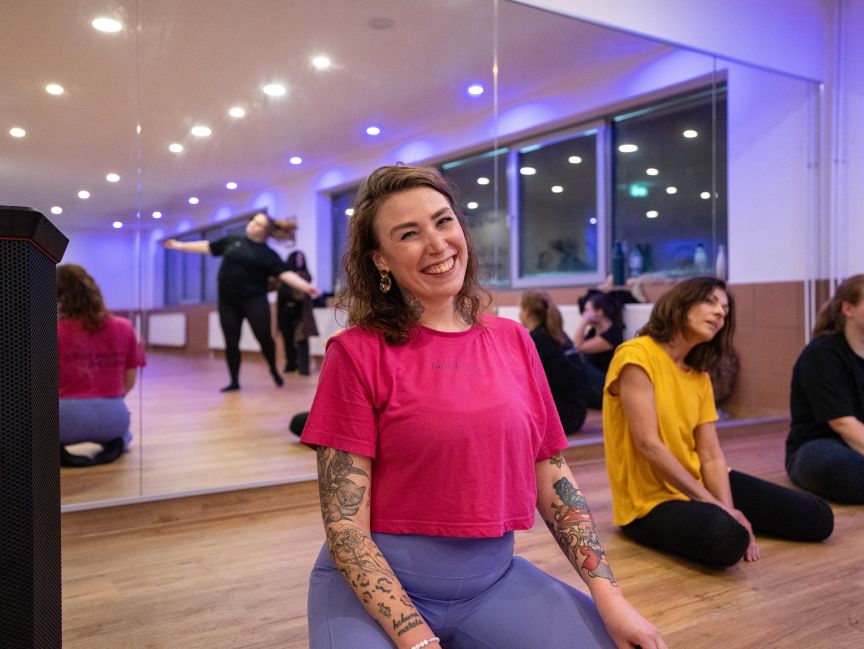 Lächelnde Frau mit Tattoos in pinkem T-Shirt sitzt in einem Tanzstudio mit Spiegelwand und anderen Personen.