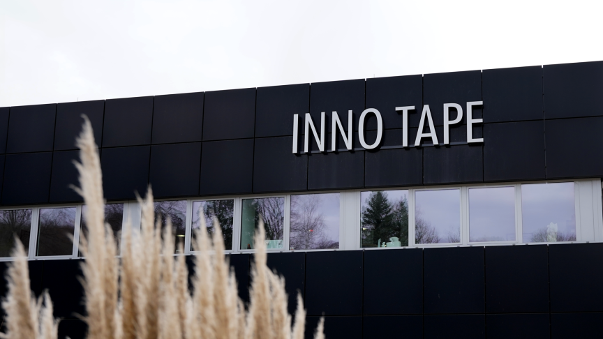 Schwarzes Firmengebäude mit weißem Schriftzug „INNO TAPE", davor verschwommene Gräser, bewölkter Himmel.