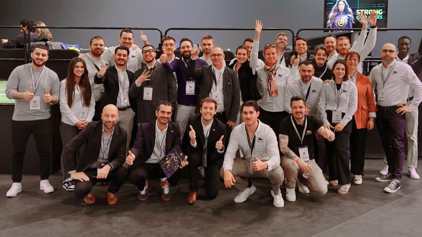 Gruppe von etwa 30 Personen mit Namensschildern posiert lächelnd für ein Gruppenfoto in einem modernen Innenraum.