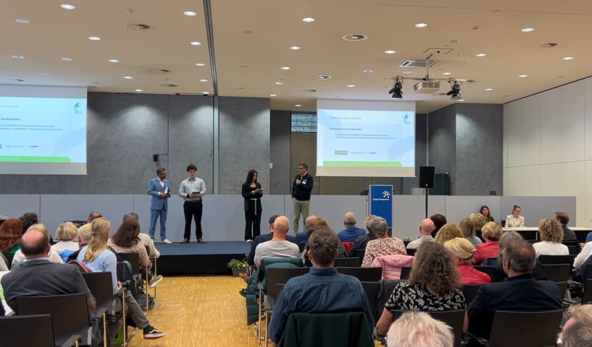 Vier Personen auf einer Bühne vor Publikum in einem Konferenzsaal mit zwei Projektionsleinwänden.