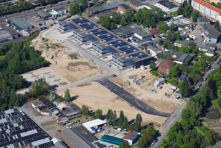 Luftaufnahme eines Industriegebiets mit Baustelle, Sandgelände, Flachdachgebäuden mit Solaranlagen und umliegender Bebauung.