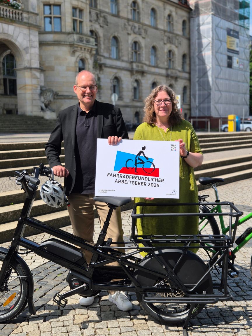 Zwei Personen halten Schild „Fahrradfreundlicher Arbeitgeber 2025" vor Gebäude, daneben zwei Fahrräder.